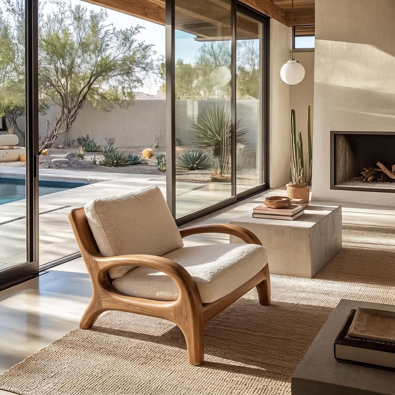A mid-century modern wooden armchair with cream bouclé cushions sits in a sunlit contemporary living room with large floor-to-ceiling windows, a concrete side table, a fireplace, and a pool visible outside.