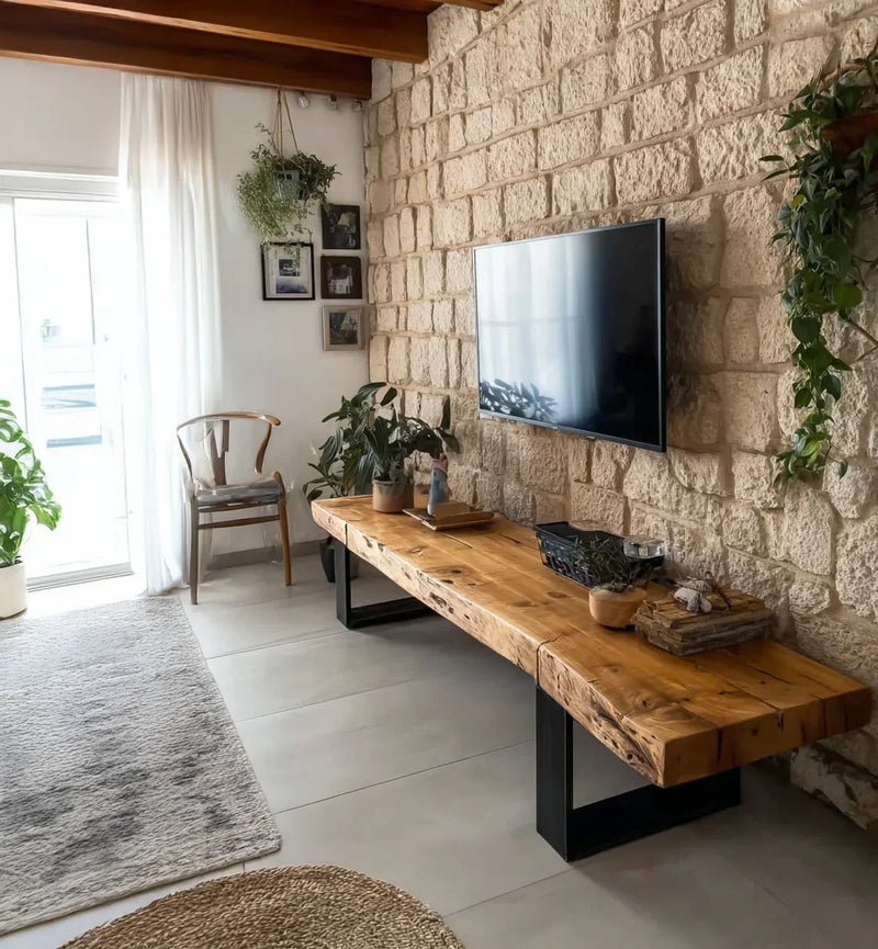 Modern living room with stone wall, wall-mounted TV, wooden bench, plants, and natural light