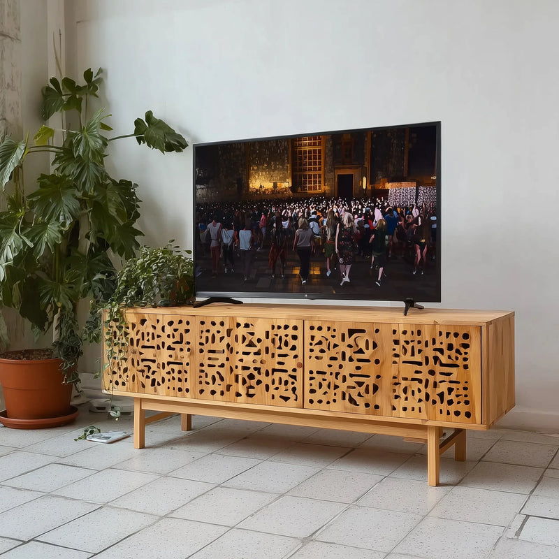 Solid wood TV stand with geometric cutout doors in a modern living room next to indoor plants.