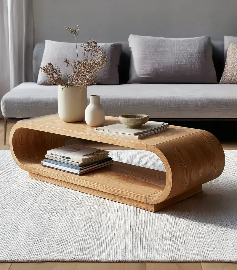 Modern living room with curved wooden coffee table, dried floral vases, books, and gray sofa