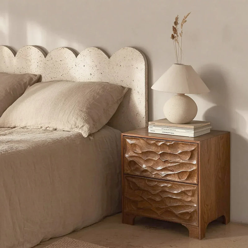 Solid wood carved nightstand with sculpted drawers beside a beige bed and textured lamp