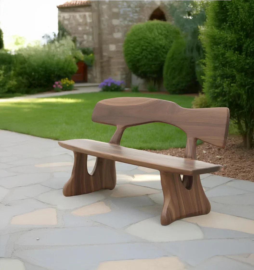 Modern wooden bench with curved design on stone patio in lush garden near stone building