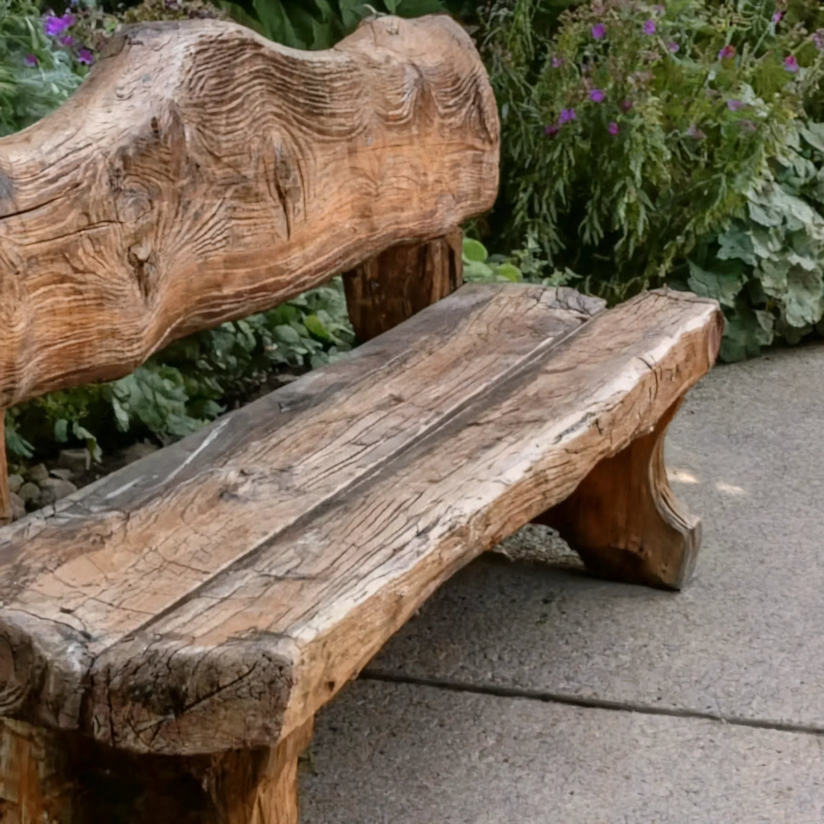 Rustic wooden bench with a carved backrest on a stone path surrounded by green plants with purple flowers