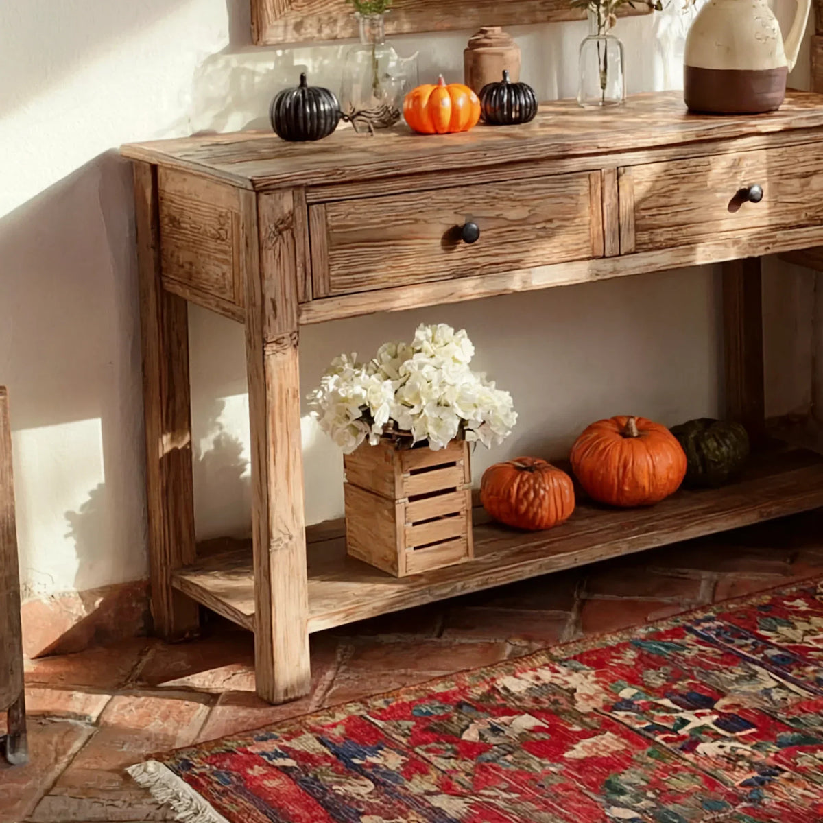 Rustic wooden console table with two drawers, pumpkins, and decorative flowers on brick flooring.