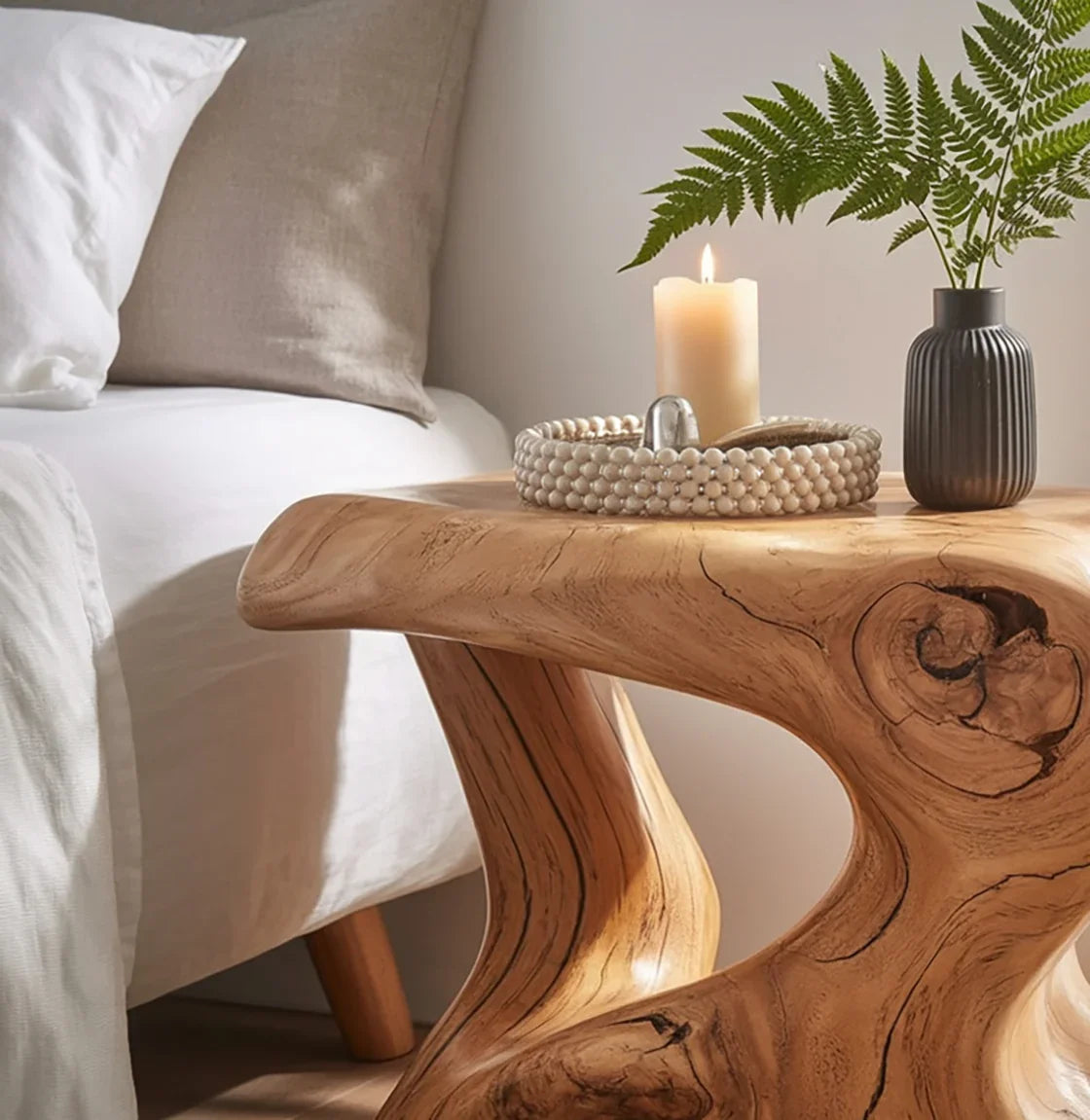 Wooden bedside table with burning candle, beaded tray, and fern in black vase next to bed