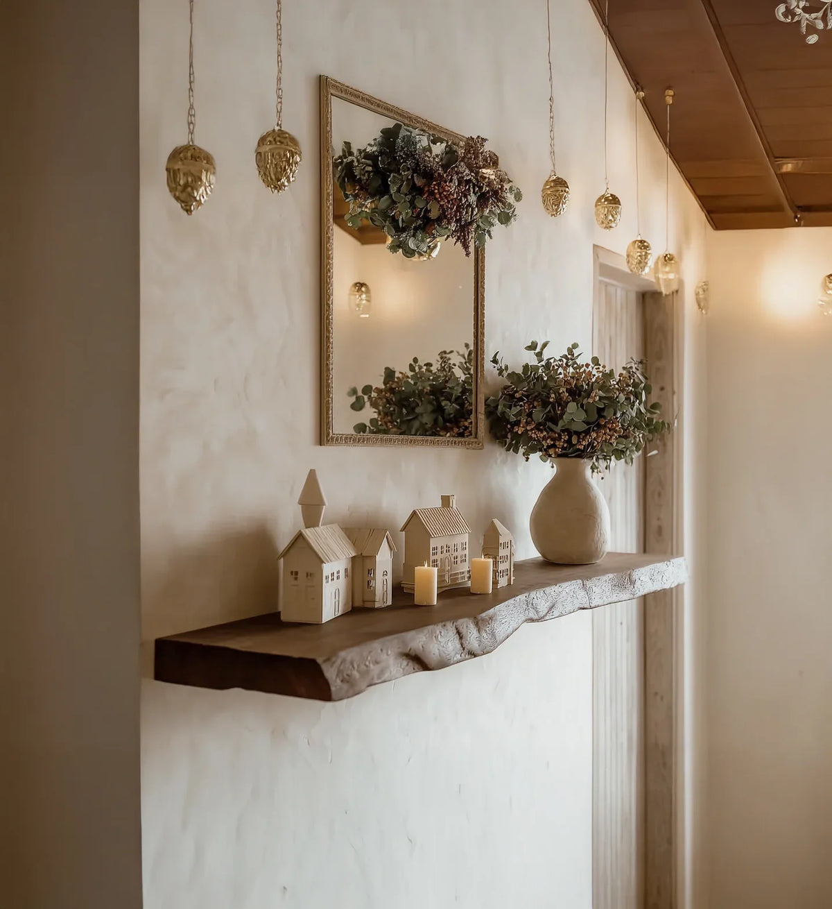 Rustic wooden shelf with decorative miniature houses, candles, and vase with greenery under hanging gold lights