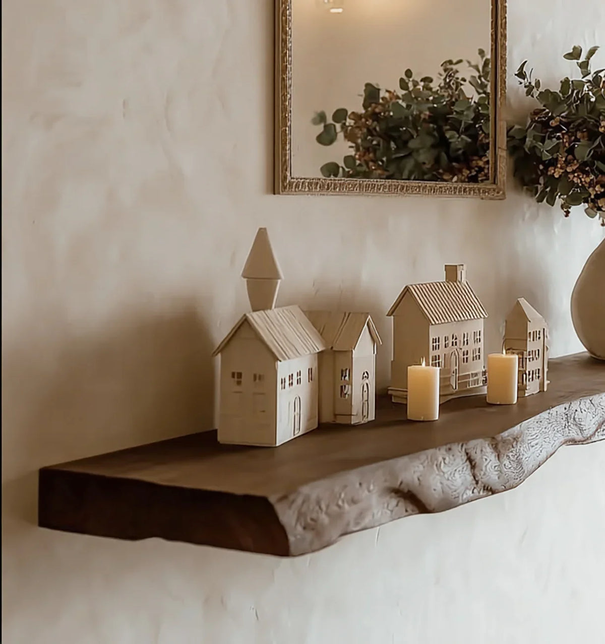 Rustic wooden shelf with miniature ceramic village houses, candles, and a mirror reflecting greenery