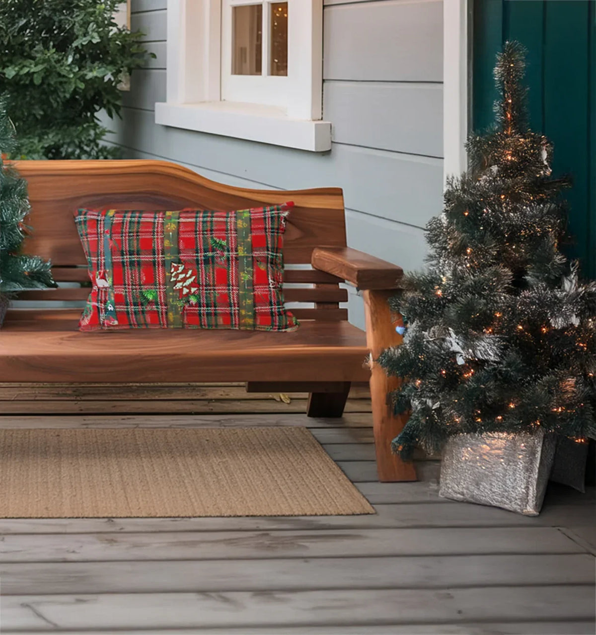 Wooden porch bench with red plaid Christmas pillow next to small decorated lit Christmas tree