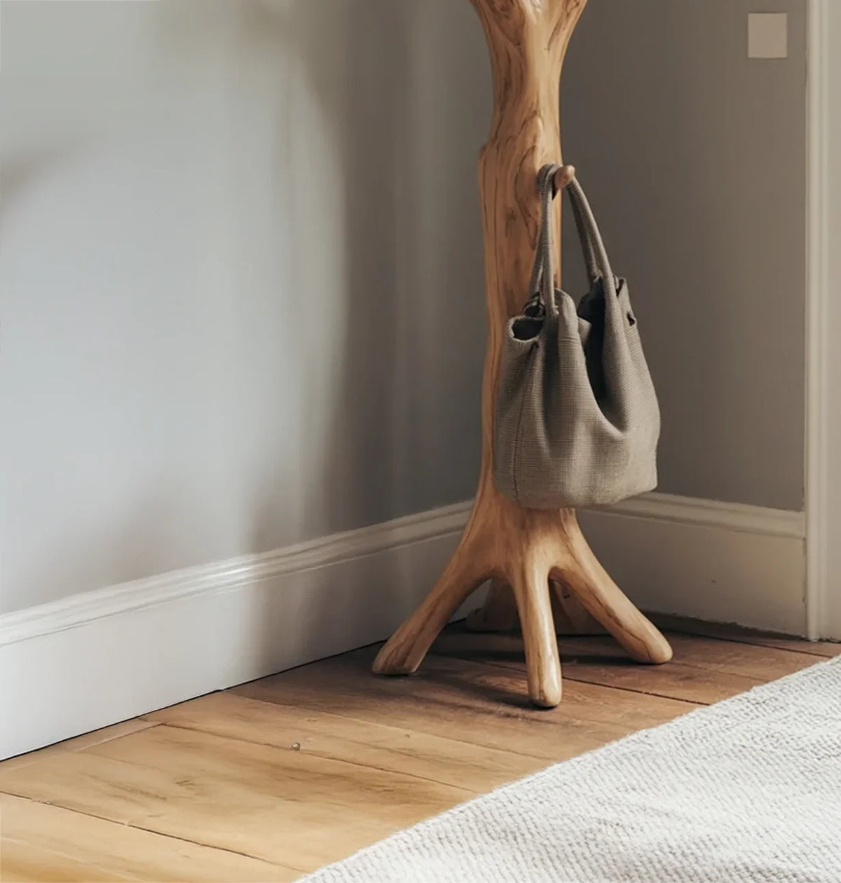Modern wooden coat rack in corner with gray textured tote bag on wooden floor and white rug