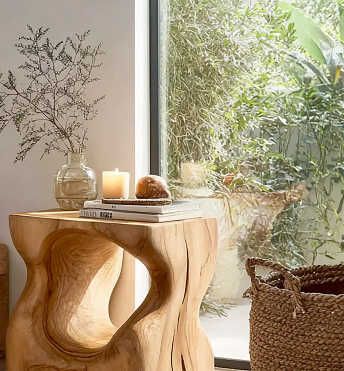 Wooden side table with dried flowers, lit candle, bread, and books near a large window with green plants