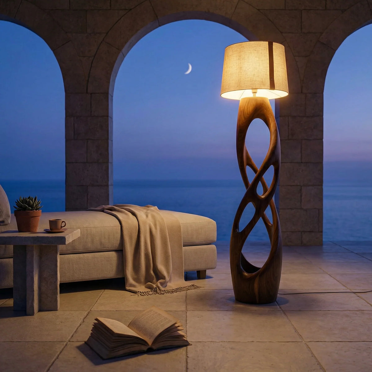 Mediterranean villa terrace at dusk with sculpted wood floor lamp, lounge chair, open book, and sea view.
