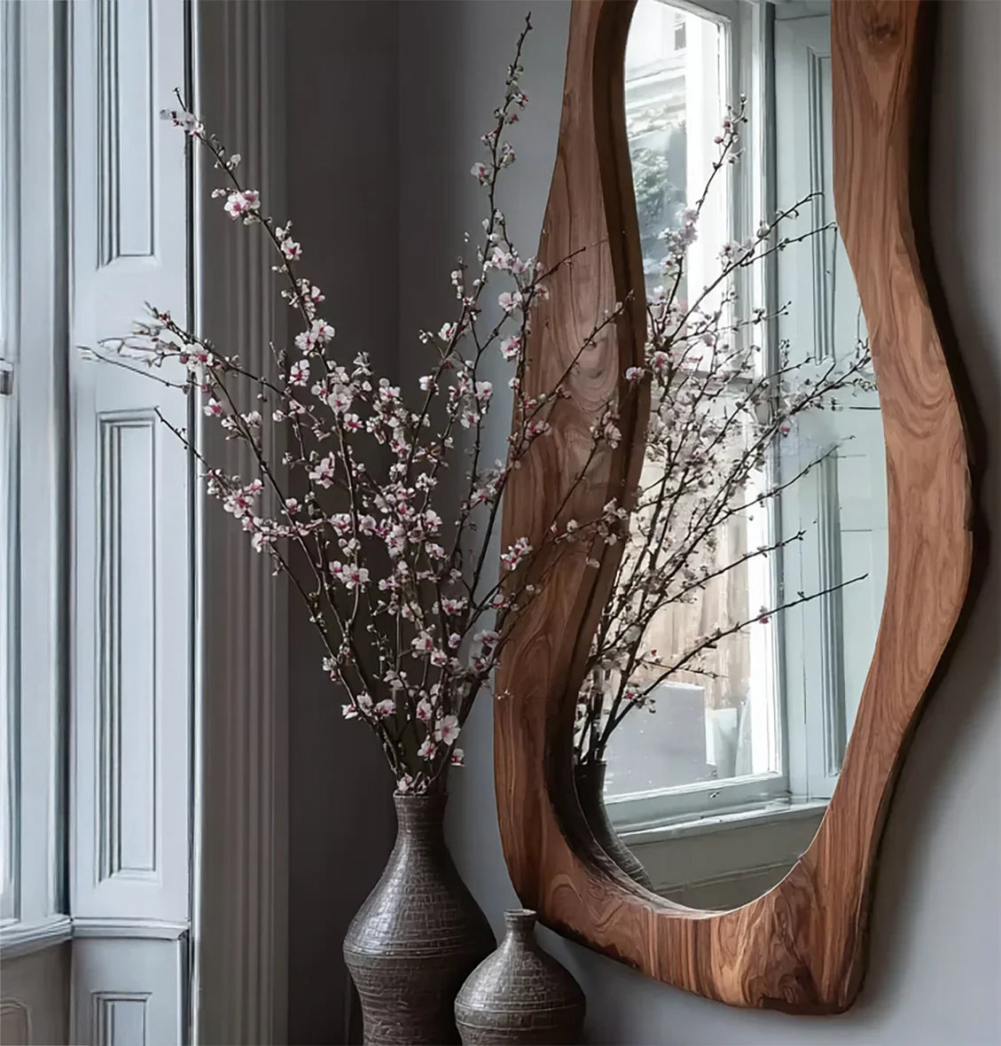 Live edge wood mirror on wall beside window with tall vases and blooming branches