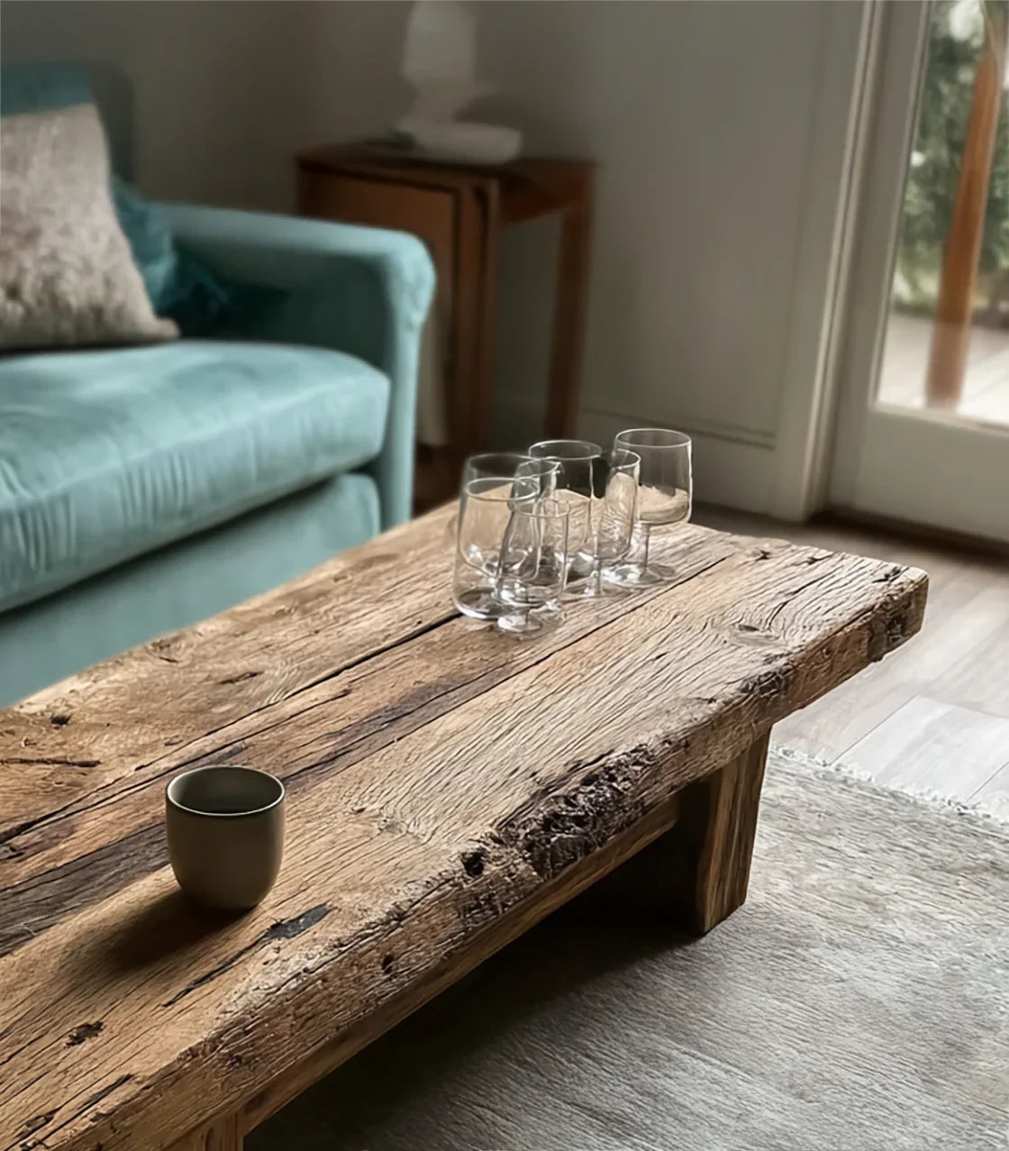 Rustic wooden coffee table with four clear glasses and a ceramic cup beside teal velvet sofa in cozy living room