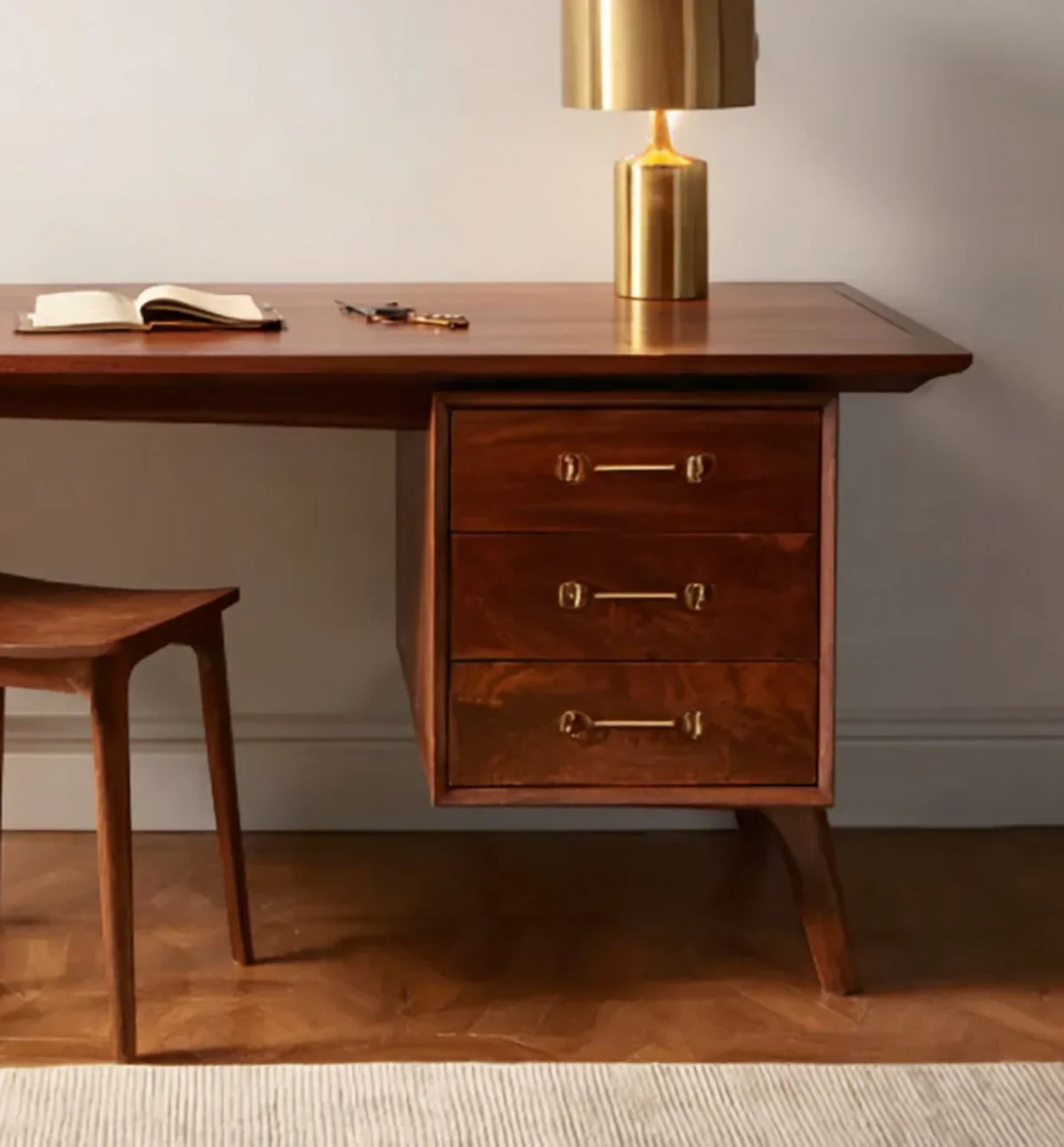Mid-century modern wooden desk with three drawers, brass handles, a gold lamp, open book, and wooden chair