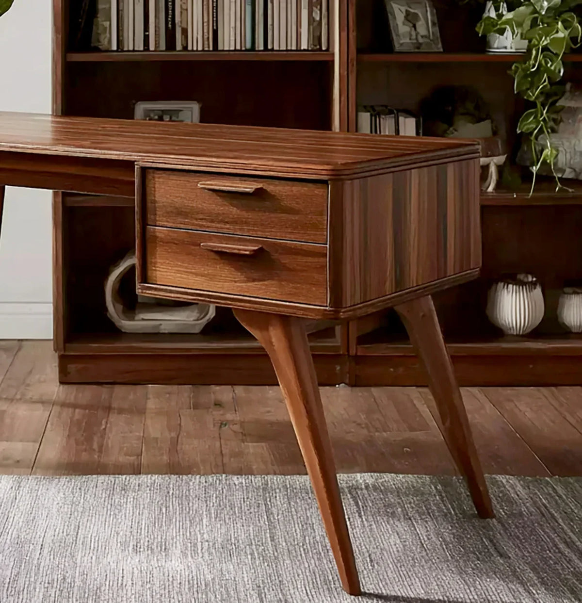 Mid-century modern wooden desk with two drawers, angled legs, and bookshelves in background