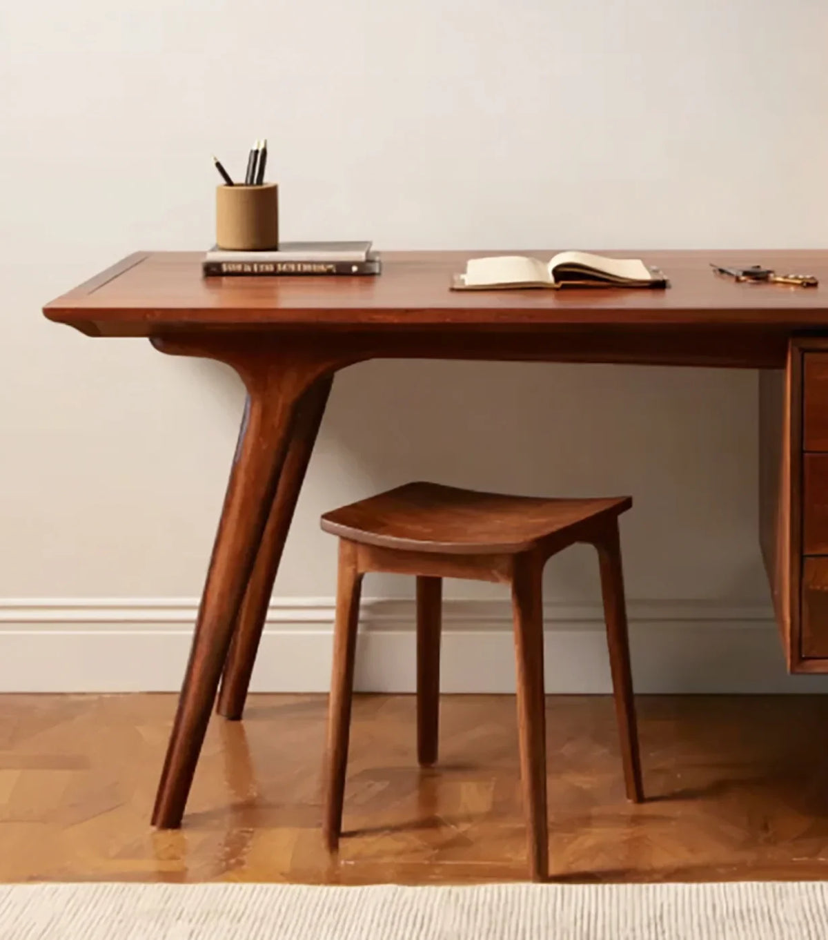 Mid-century modern wooden desk with matching stool, book, pencil holder, and keys on hardwood floor