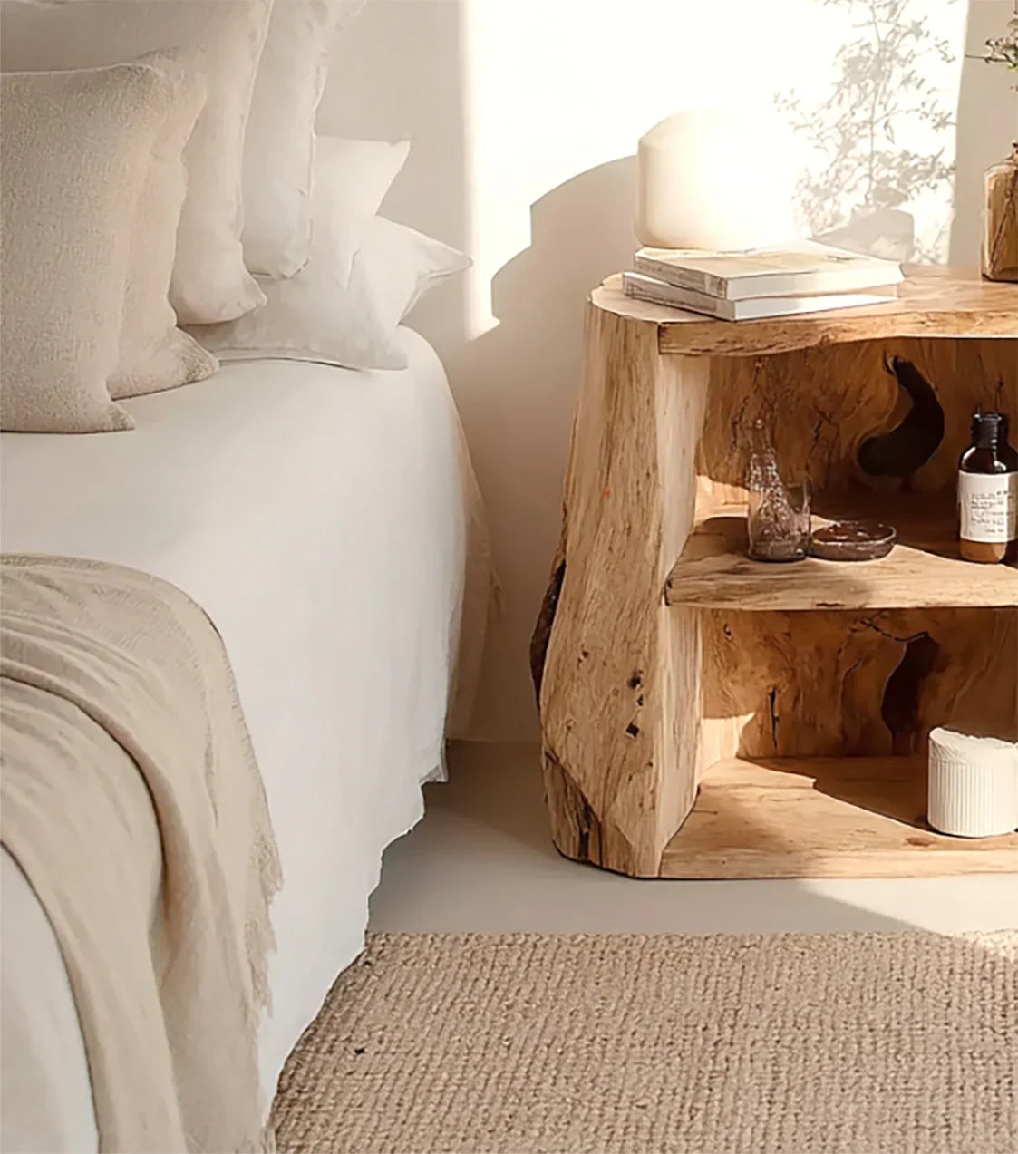 Cozy bedroom corner with white bed linens, beige pillows, woven rug, and rustic wooden nightstand
