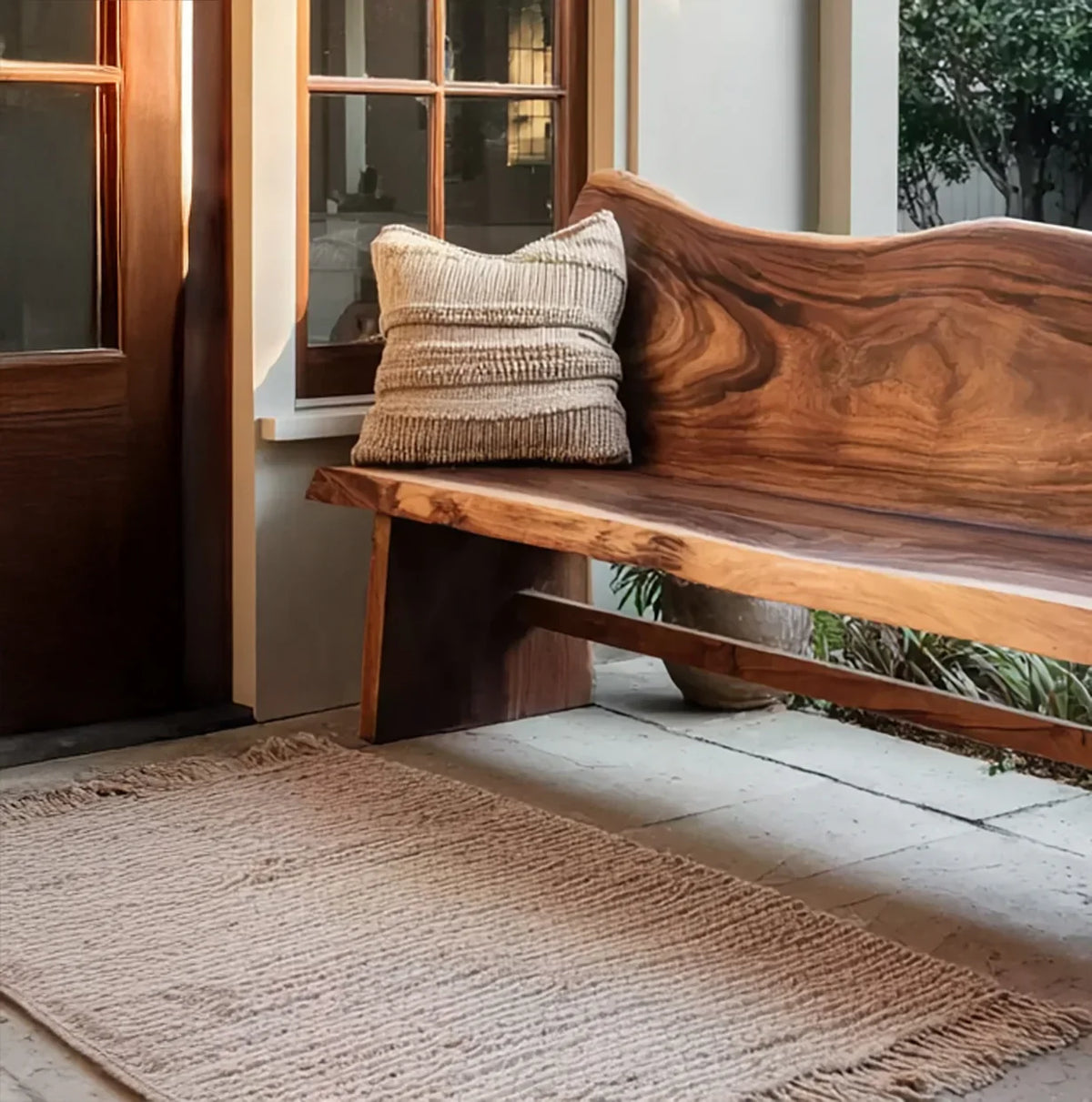 Natural wood bench with textured pillow by window on stone floor with woven rug