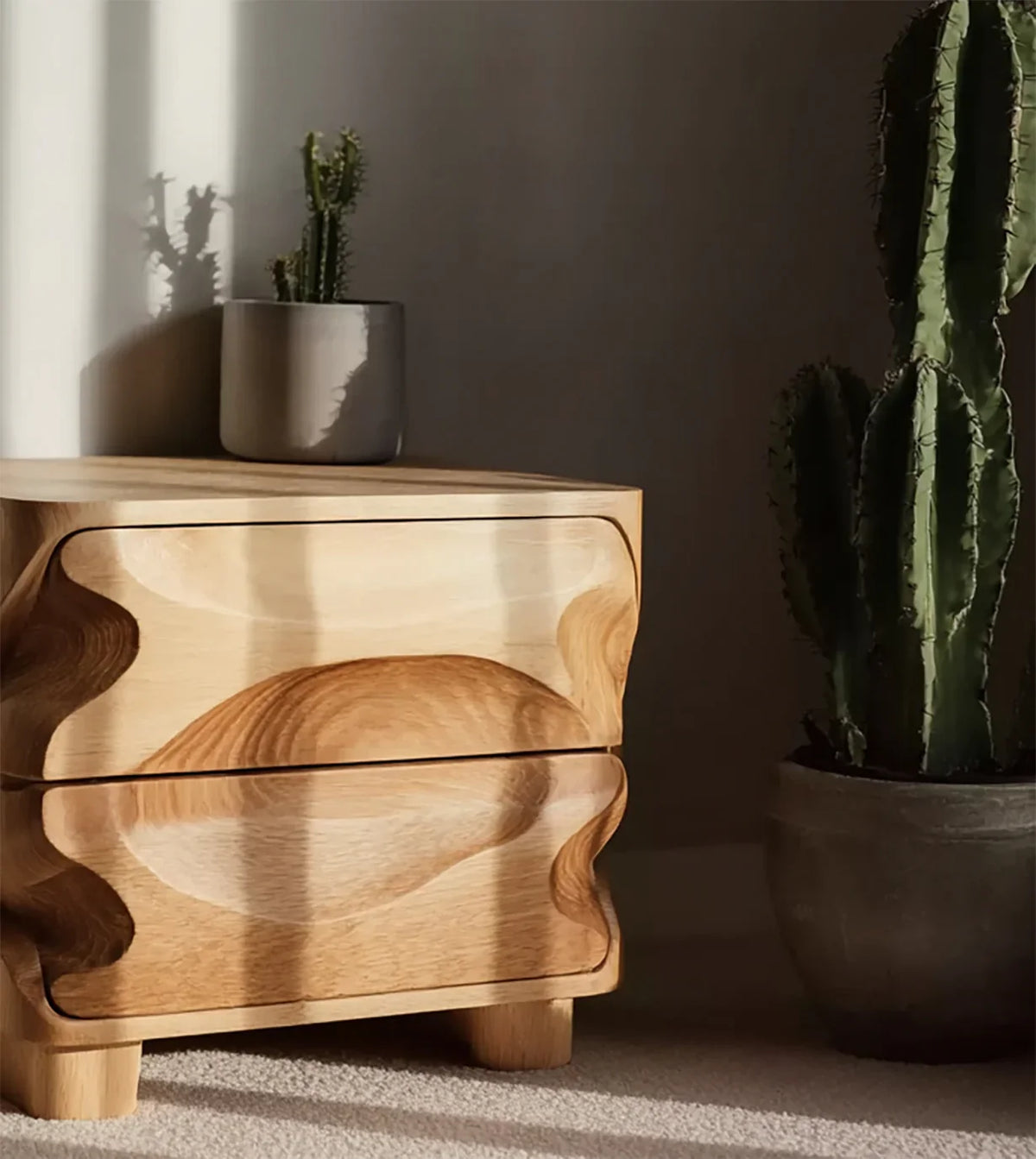 Wooden nightstand with sculptural drawers next to potted tall cactus and small cactus in gray pot