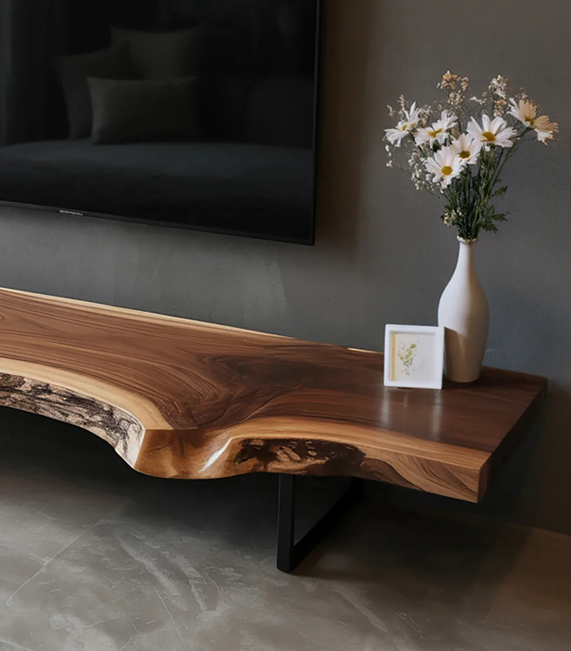 Live edge wooden TV console with white vase of daisies and framed art on gray wall and concrete floor