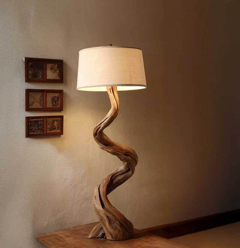 Twisted wooden floor lamp with white fabric shade on wooden table against beige wall with small framed pictures