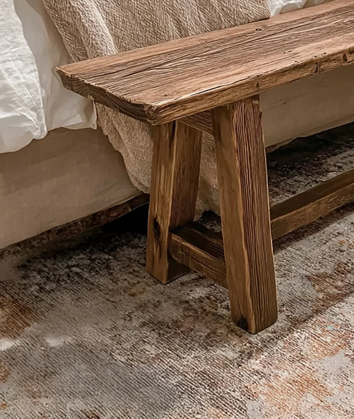 Rustic wooden bench with natural grain, next to bed with white and beige linens on textured rug