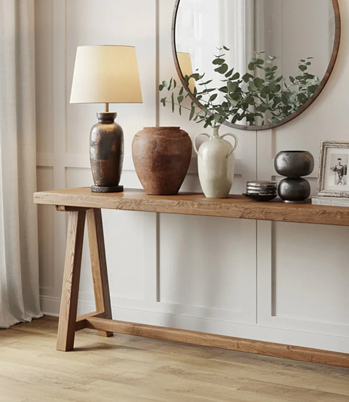 Wooden console table with a table lamp, ceramic vases, greenery, and round wall mirror in beige room