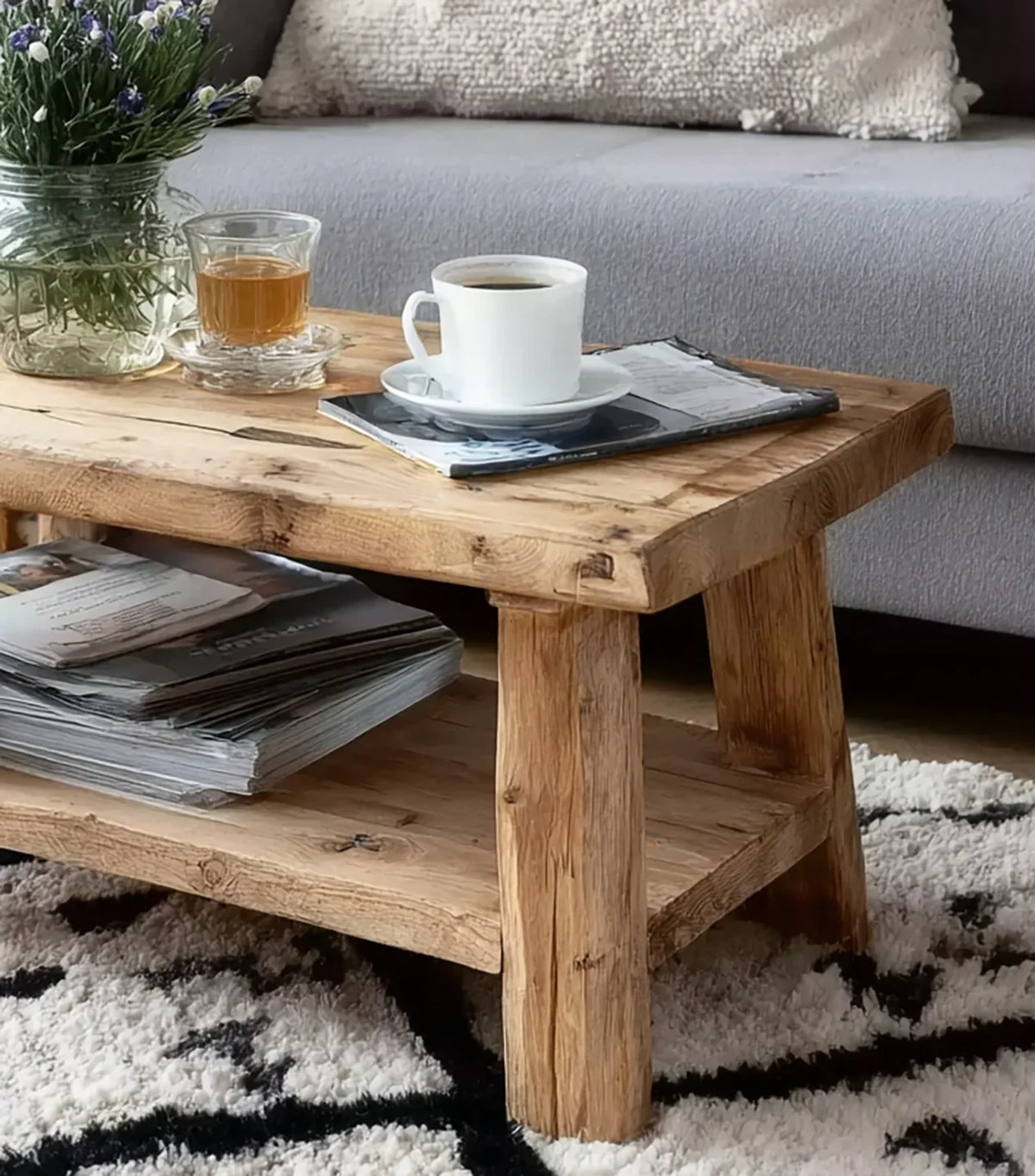Rustic wooden coffee table with coffee cup, glass of tea, magazines, and flower vase on a patterned rug