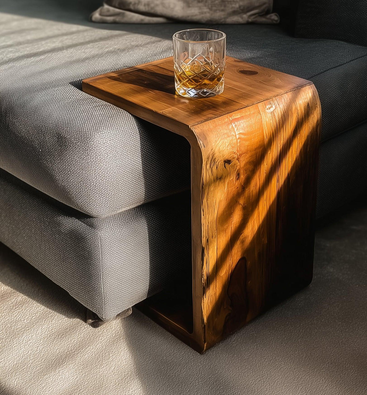 Modern wooden side table over gray sofa armrest with glass of whiskey in natural sunlight