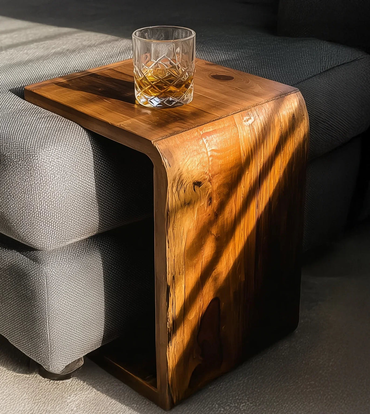 Natural wood side table on gray fabric sofa with glass of whiskey, warm sunlight and shadows