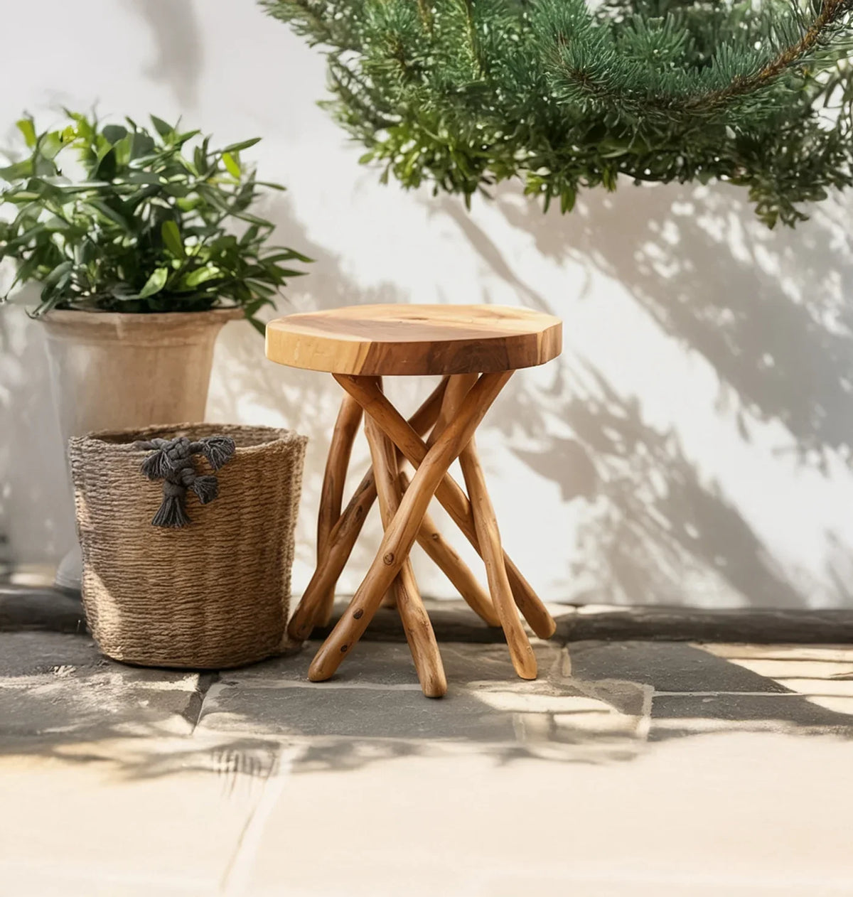 Natural wood side table with crossed legs on stone floor next to woven basket and green plants