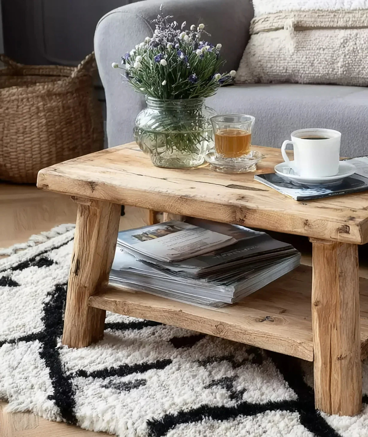 Rustic wooden coffee table with glass vase flowers, tea, coffee cup, magazines, on patterned rug near gray sofa