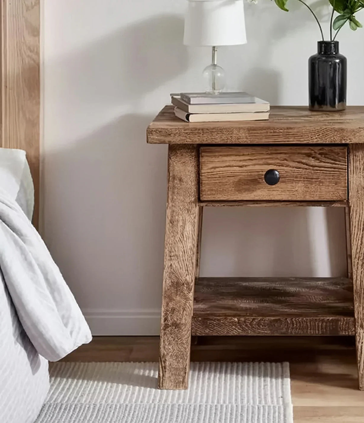Rustic wooden nightstand with drawer, stacked books, white lamp, and black vase with flowers