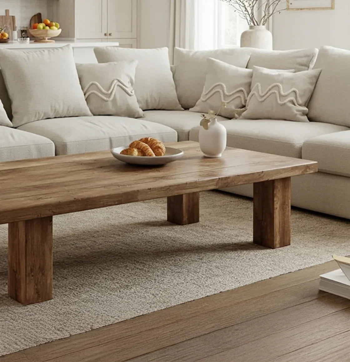 Modern living room with beige sectional sofa, textured pillows, wooden coffee table, croissants, and minimalist decor