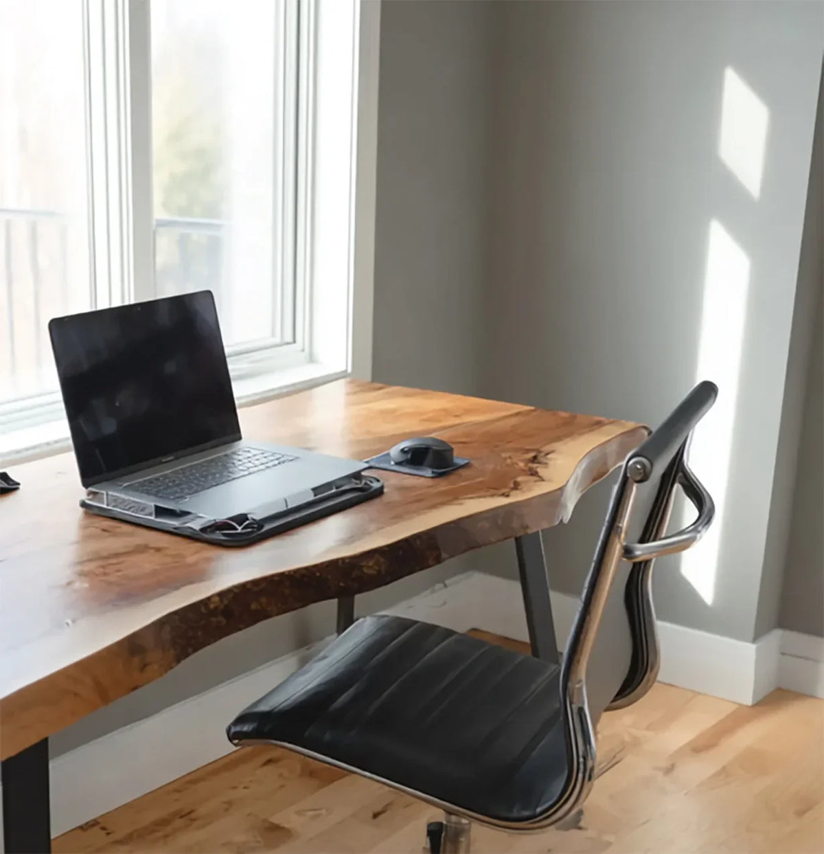 Modern home office with wooden live-edge desk, black leather chair, laptop, and mouse by window