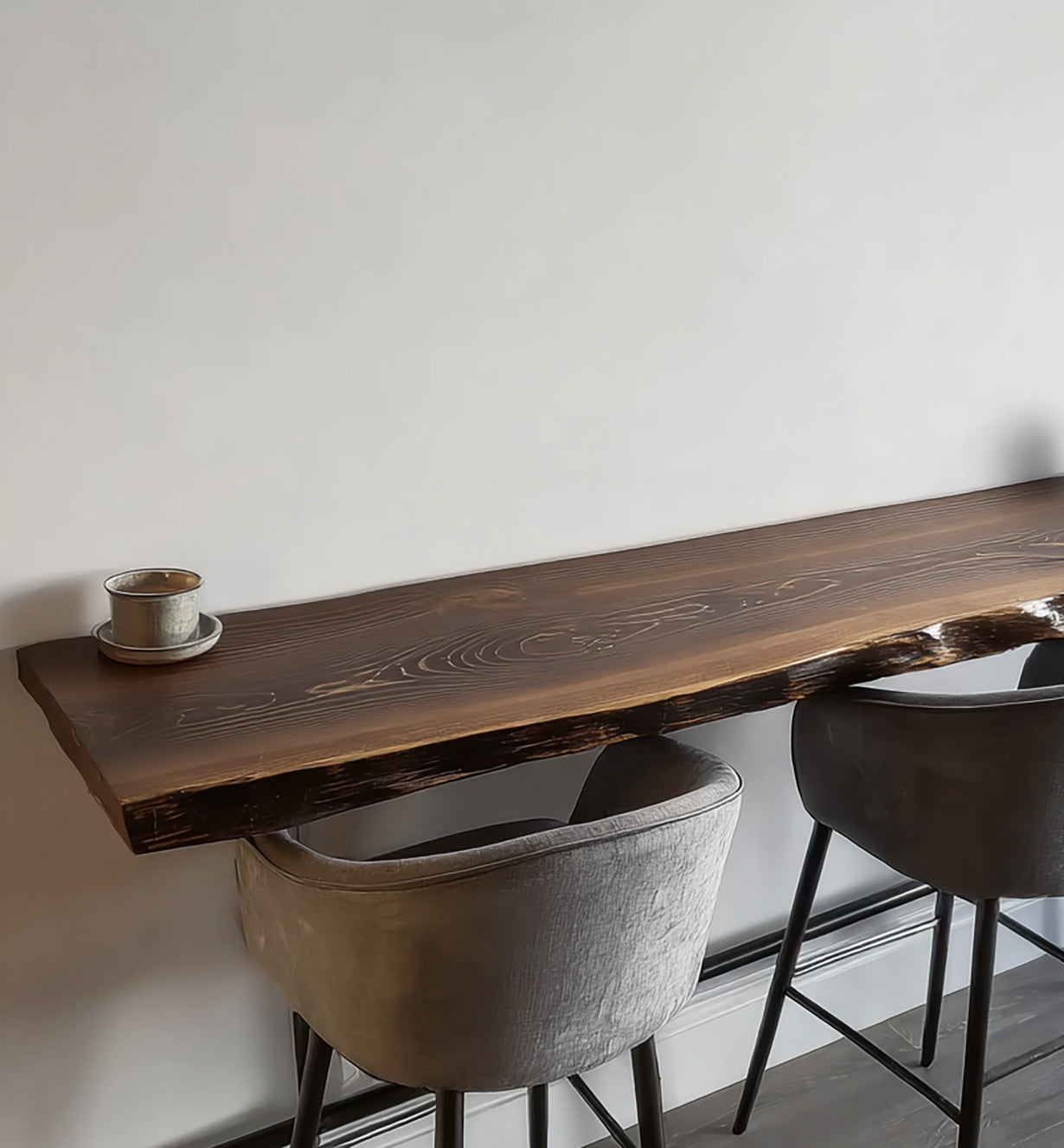 Long dark wooden bar table with live edge and two gray velvet bar stools against white wall