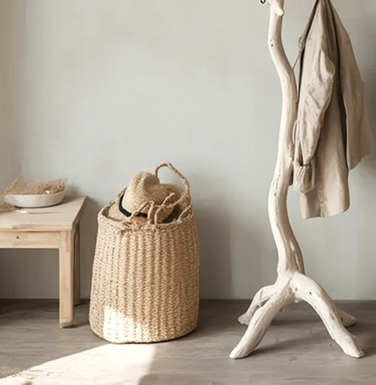 Woven basket with straw hat and bag beside wooden bench and white driftwood coat rack with beige jacket